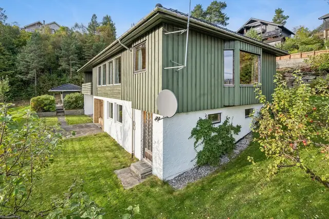 Boligen følger det skrånende terrenget, med hage langs fasaden og vinduer i begge etasjer.